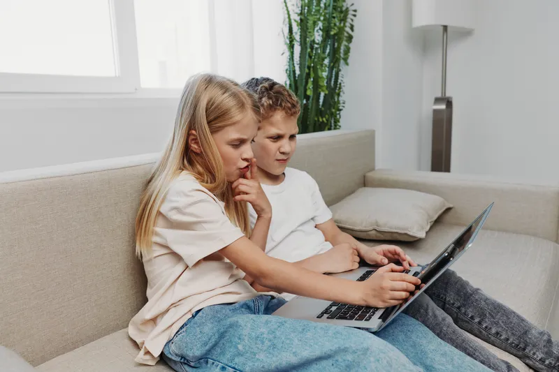 Two focused children sitting on a beige sofa using a laptop together in a bright living room. Concept of learning, technology, childhood, cooperation, and digital education.
