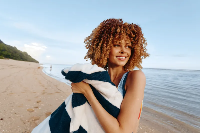 Cheerful woman with curly hair on a sunny beach, holding a striped towel, smiling toward camera, coastal ocean view behind her, relaxed summer mood, soft sea breeze natural light and sand