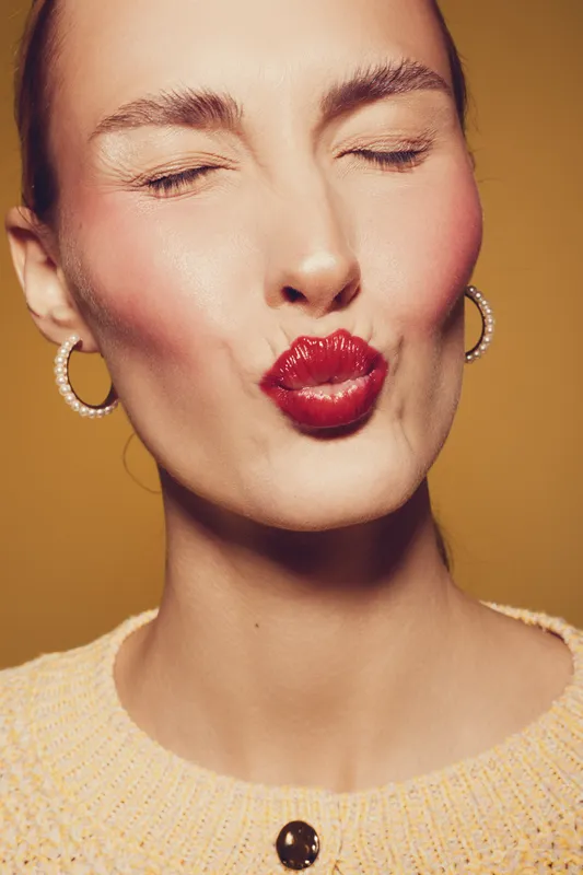 Close-up of a woman with red lipstick puckering lips and closed eyes. She wears hoop earrings and a knitted sweater. Concept of beauty, makeup, and facial expression.