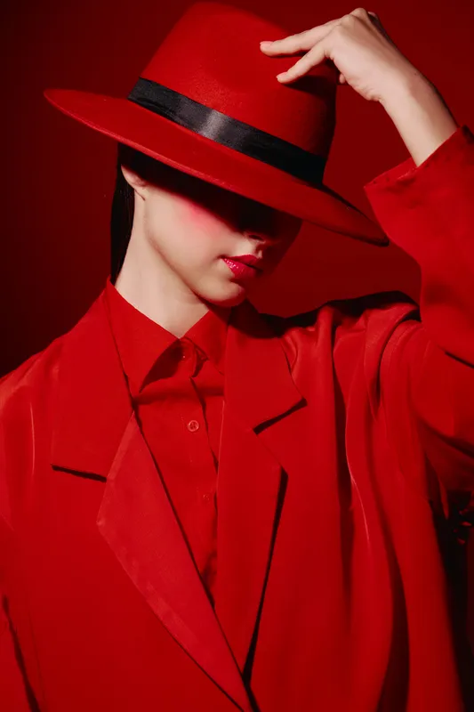 Fashion studio portrait of a stylish woman wearing a red hat and oversized red suit, face partially hidden. Creative beauty concept with vibrant red tones and dramatic shadows.