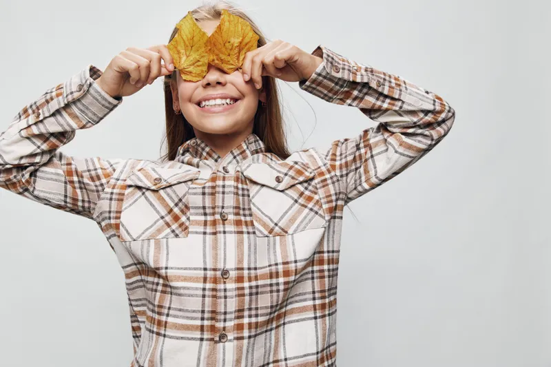 Girl smiling and holding autumn leaves over eyes wearing checkered shirt, isolated on white background. Child playing with nature elements and showing happy expression.