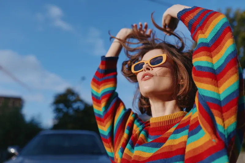 Woman in sunglasses and colorful sweater posing outdoors in a fashion portrait, enjoying sunlight and breeze with hands in hair, street style vibe and vibrant casual look.