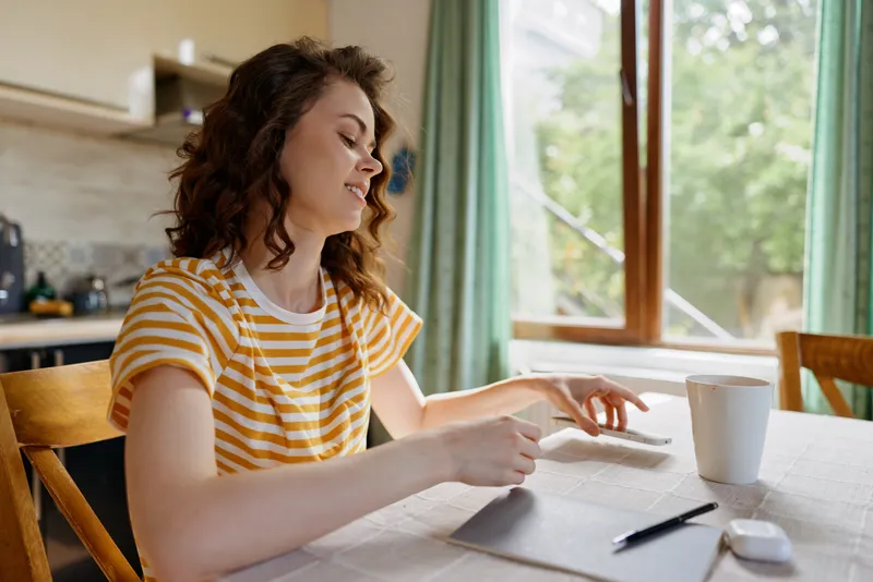 Smiling woman with wavy hair at home, seated at a sunlit dining table by a window, wearing a striped yellow shirt, writing on paper, with a coffee mug nearby, showcasing casual lifestyle and cozy