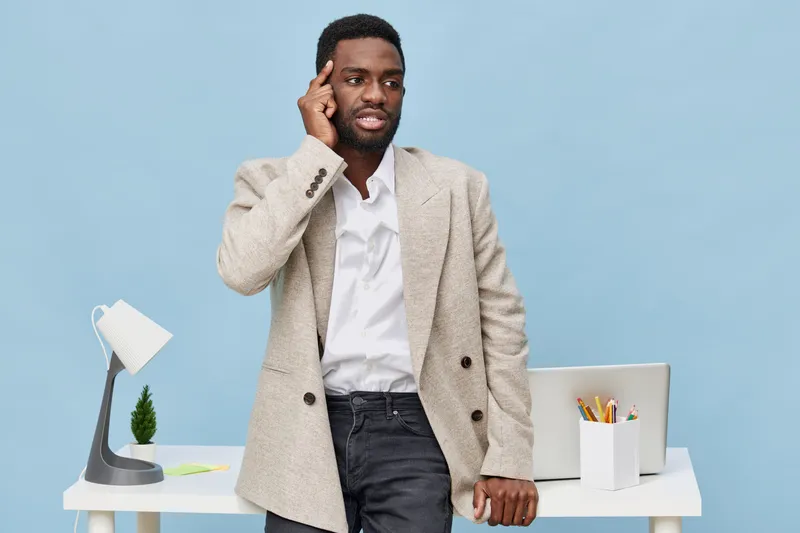 Confused man with headset in stylish beige blazer standing by desk with laptop, stationery, and lamp on light blue background. Studio shot for business, communication, or customer support concepts.