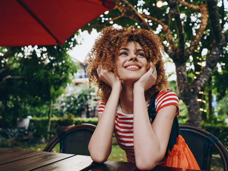 Joyful young woman with curly hair sits at an outdoor cafe table under a red umbrella, smiling with hands resting on her chin, sunlight and greenery framing her scene.