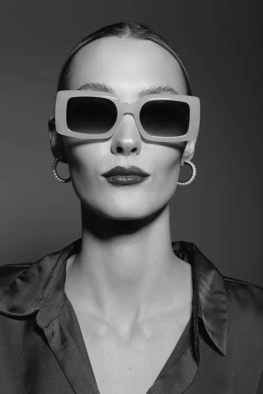 Stylish Caucasian woman with pale skin wearing large sunglasses and hoop earrings poses confidently in black and white portrait with dramatic lighting.
