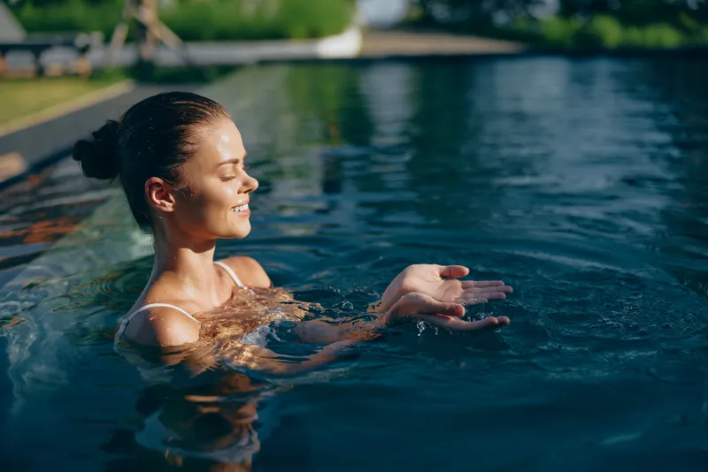 A woman enjoys a sunny pool moment, smiling as sunlight dances on blue water. A relaxed, casual scene emphasizing poolside leisure, wellness, and outdoor recreation.