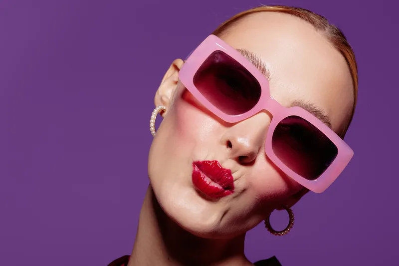Close-up of a woman wearing large pink sunglasses and red lipstick making a kiss face on purple background. Concept of fashion, beauty, and playful attitude.