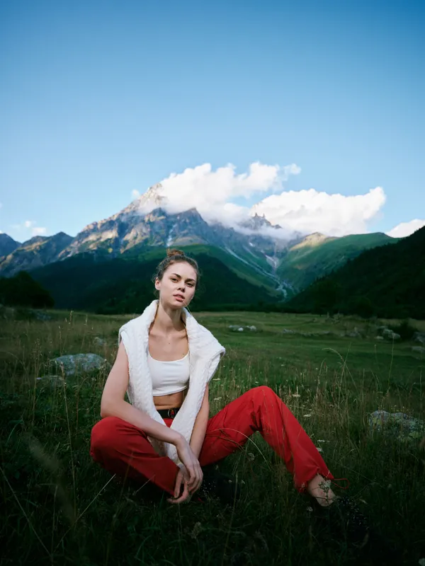 Woman in red pants and white top sits in a meadow before towering mountains, highlighting outdoor fashion, nature, and tranquil scenery.