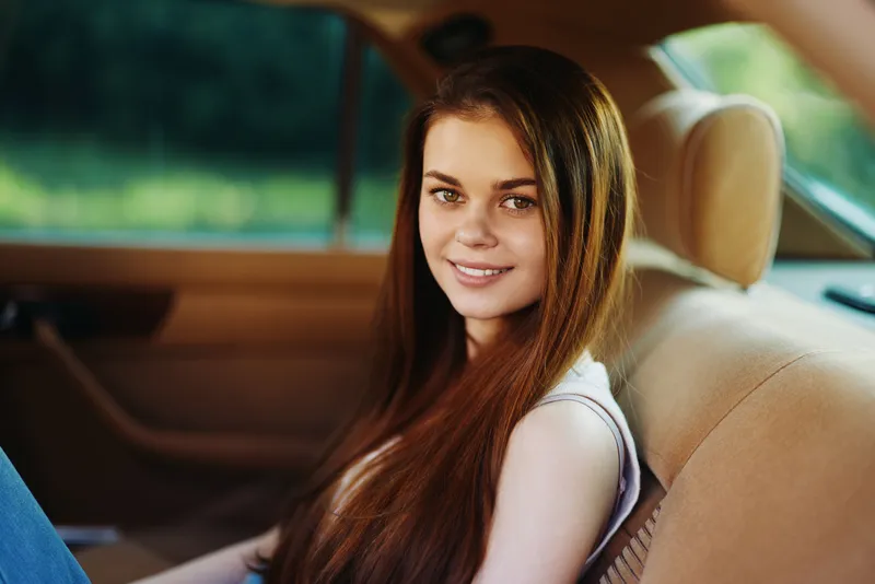 Young woman with long hair sitting in a car, smiling with a relaxed expression, showcasing casual style in a sunny outdoor setting.