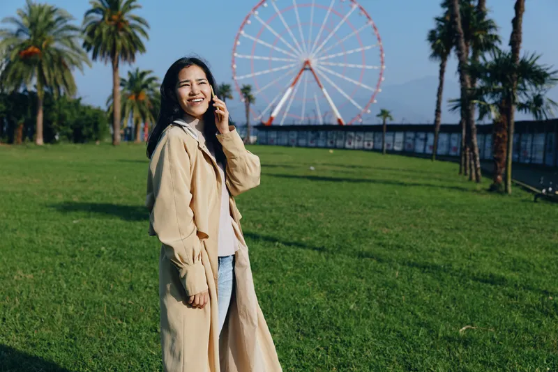 Woman in a beige coat smiles while talking on a phone in a sunny park with a ferris wheel in the background, lush grass and palm trees.