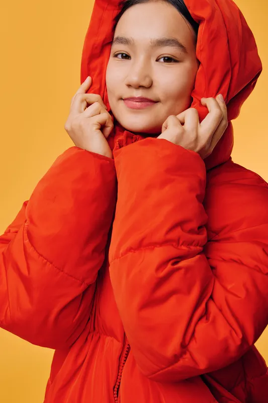 Young woman in bright red winter jacket with hood smiles gently on yellow background. Female model with natural makeup wearing warm clothing for cold weather portrait.