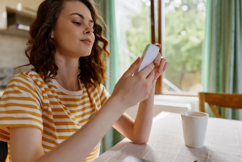 Casual woman with wavy hair using a smartphone at a bright cafe table, striped yellow and white shirt, soft natural light creates a warm, everyday lifestyle moment.