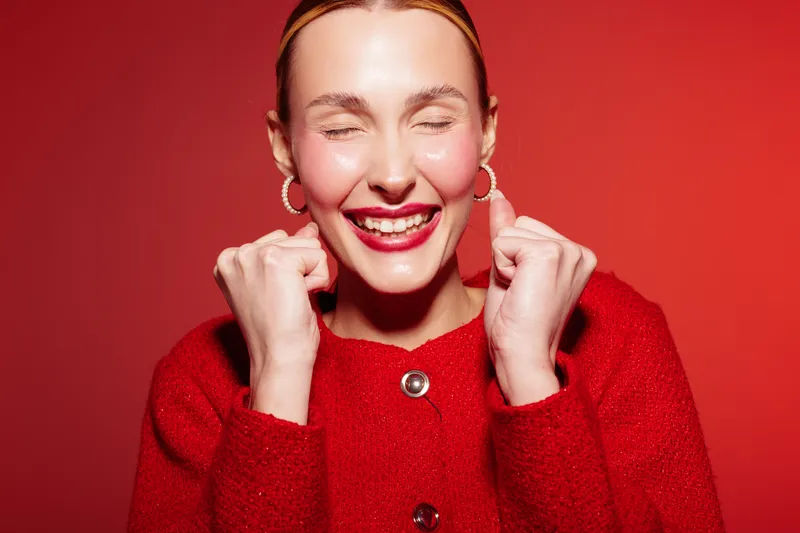 Happy young woman in red sweater smiles with closed eyes and clenched fists on red background. Concept of joy, excitement, celebration, and positive emotions in studio lighting.