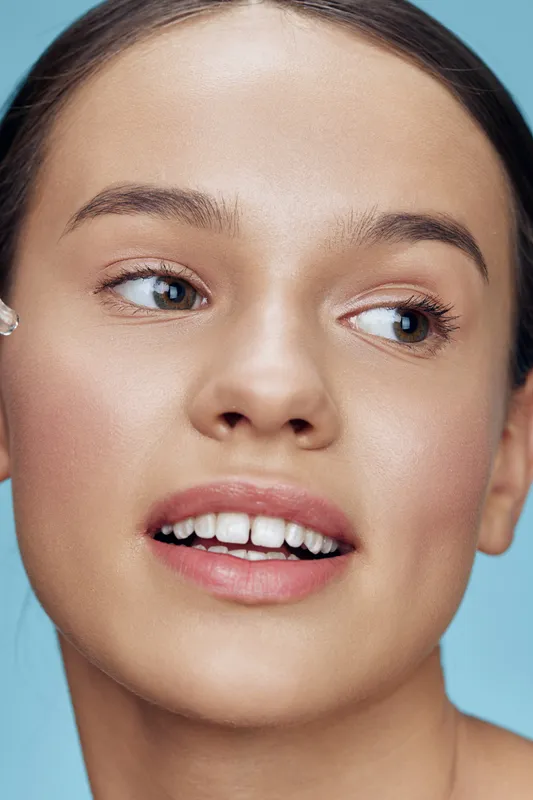 Young woman applying skincare product on her face, showcasing natural beauty and healthy skin, with a soft blue background for a fresh aesthetic