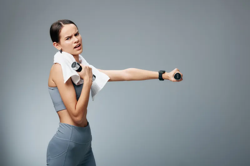 Fitness woman demonstrates workout with dumbbell, towel over shoulder, smartwatch on wrist, gym attire, studio backdrop, focused movement and athletic energy.
