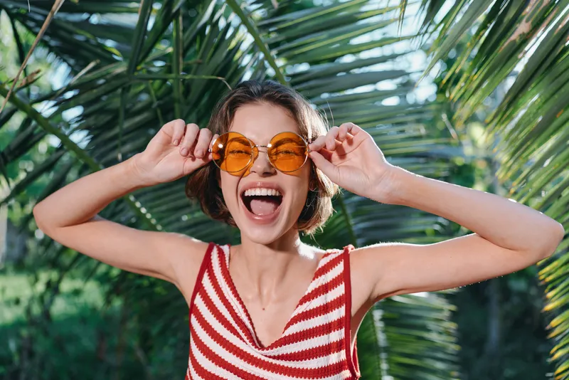 A cheerful woman wearing a red and white striped top smiles broadly while adjusting bright orange sunglasses outdoors, surrounded by lush tropical greenery, warm sunlight, and joyful breeze.