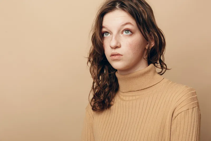 Young woman with curly brown hair wearing beige turtleneck sweater looks up thoughtfully against plain beige background. Concept of contemplation, emotion, and natural beauty portrait.