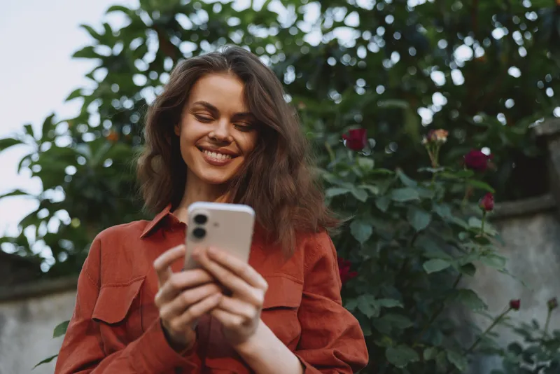 A smiling young woman with brown hair takes a casual selfie in a lush garden wearing a rust red jacket, capturing a candid lifestyle moment in natural light, ideal for social media style stock use