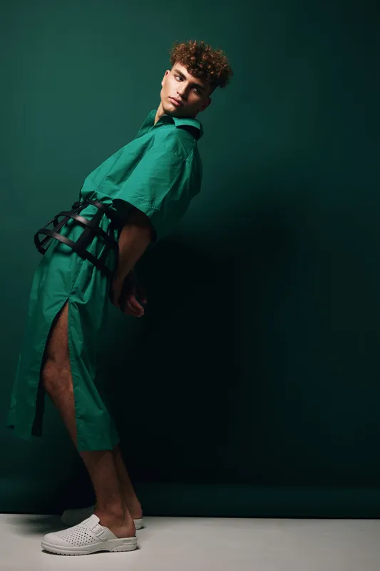 Young man with curly hair posing in teal outfit with slit pants and black belt standing sideways against green wall with shadow and white shoes in studio light.