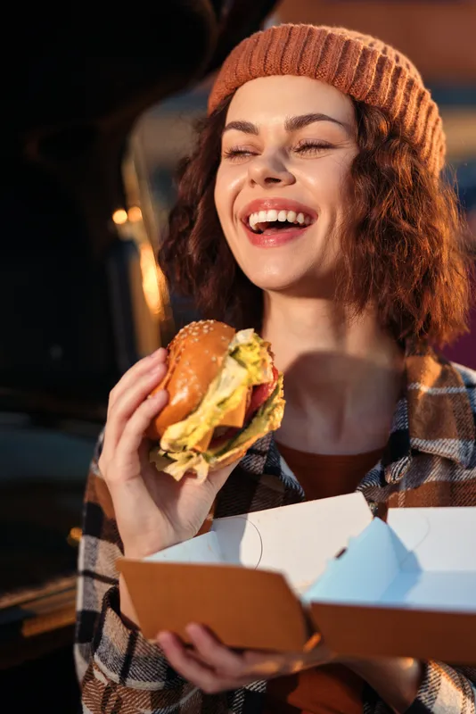 Woman holding burger and takeaway box, smile and laugh in candid moment with golden glow, authenticity and lifestyle vibe, mindful living and emotional storytelling in urban evening.