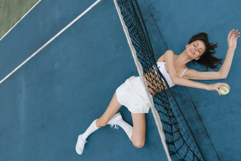 Young female athlete enjoying a tennis match on a blue court, wearing a sporty outfit, smiling with a tennis ball in hand, embodying energy and fun