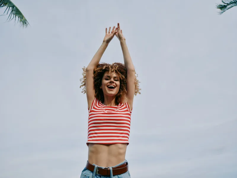 Vibrant portrait of a joyful woman raising arms beneath a clear sky, wearing a red and white striped top and denim jeans, radiating carefree summer energy.