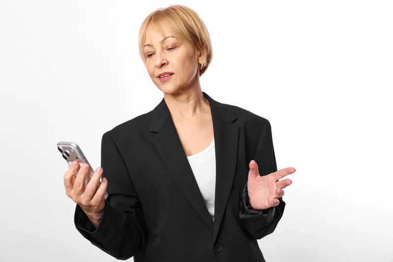 Mature woman in business suit holds a smartphone for communication gesture with mobile work on a white background Clean studio lighting shows confidence