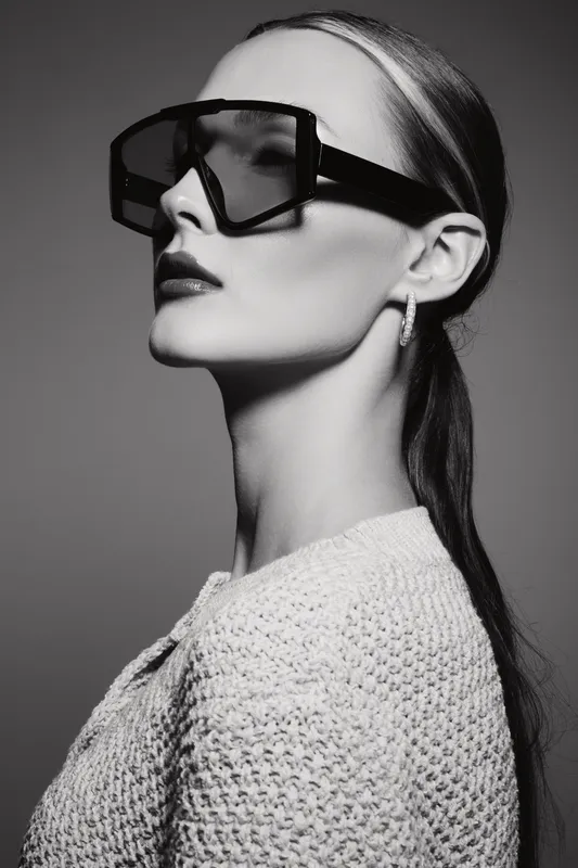 Elegant white woman with pale skin wears large modern sunglasses and knitted sweater. Studio portrait in black and white with high contrast lighting.