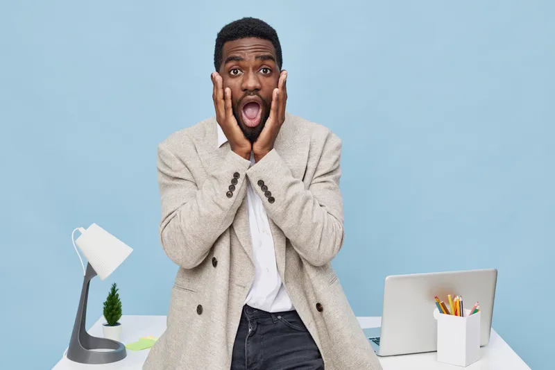 Frightened man with surprised expression holding face with hands in office environment, feeling shocked or scared, with computer and desk, light blue background, studio shot, lifestyle concept, emotio