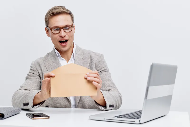 Happy surprised young man opening envelope in office at desk with laptop, smartphone, calculator. Casual business attire, light background. Concept: receiving good news, success, achievement, career, 