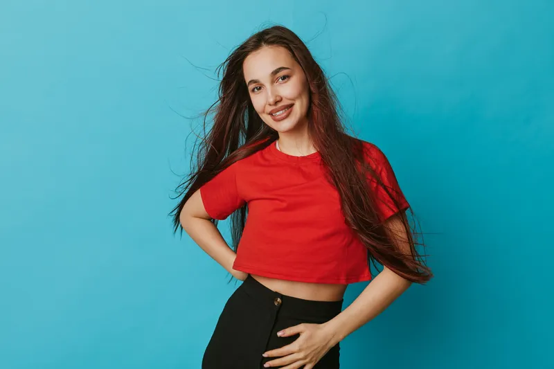 Young woman with long hair wearing red crop top and black pants posing with hand on hip smiling against blue background. Casual style, confident and happy female portrait.