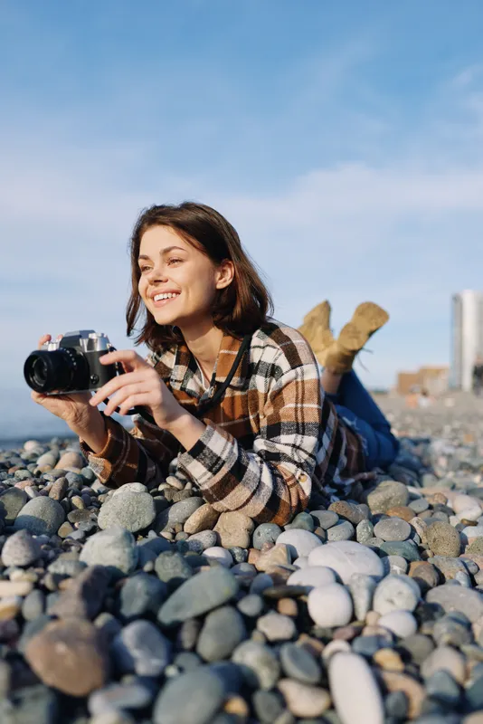 Woman photographer lying among smooth pebbles on a sunny beach with a camera, capturing a candid moment in outdoor lifestyle photography near the coast and sea horizon