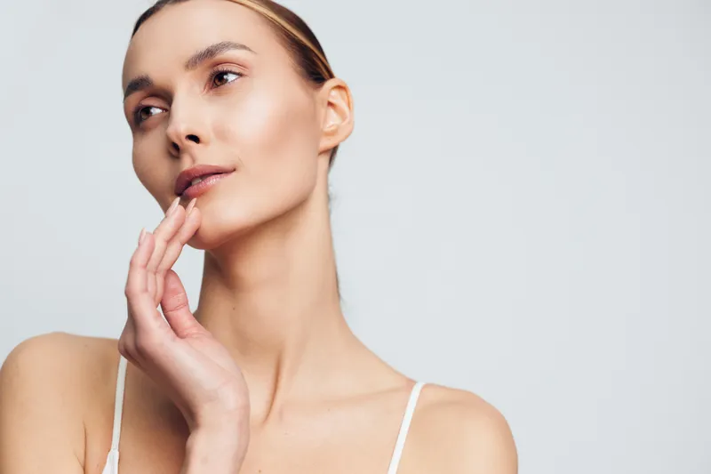 A young woman with clear skin and natural makeup gently touches her face, looking thoughtful against a plain light background. Concept for skincare, beauty, and self care.