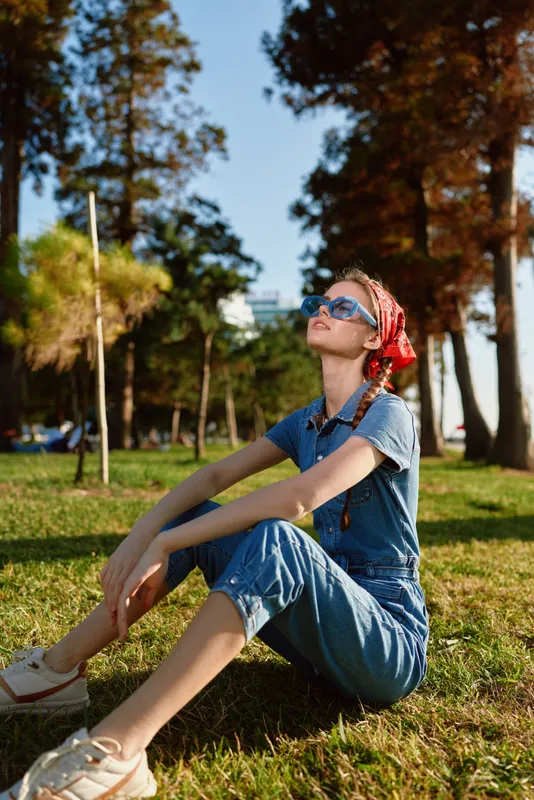 Lifestyle woman in modern boho-western denim street style sits relaxed on grassy park lawn with film color effect, wearing sunglasses and a red headscarf on sunny day.
