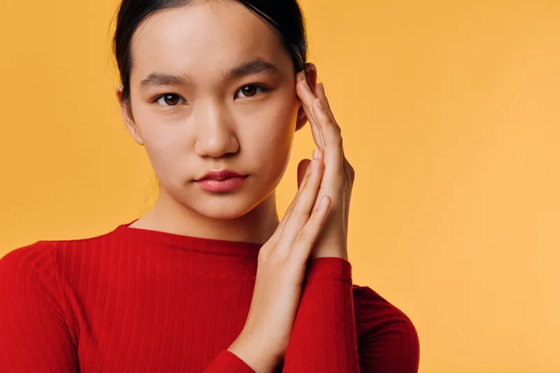 Young Asian woman wearing red sweater posing with hands near face against orange background. Female model with serious expression, smooth skin and dark hair in simple studio portrait.