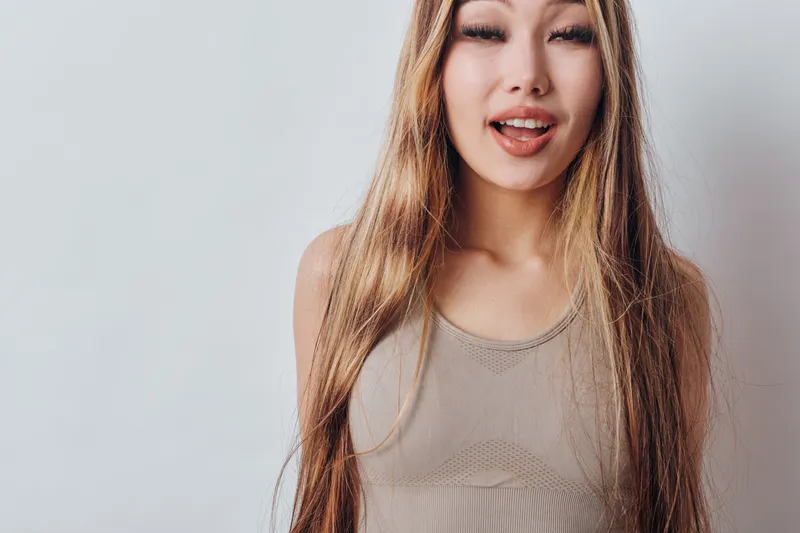 Young woman with long hair wearing casual top winking and sticking out tongue on neutral background studio portrait for lifestyle and emotion concept.