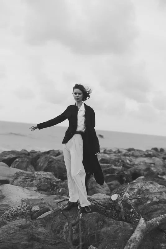 Fashion model stands on rugged rocks by the sea, blazer over white pants, wind in hair, a monochrome outdoor moment.