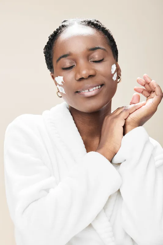 Black woman with braids smiling while applying facial cream, wearing a white bathrobe on a soft beige background, embodying skincare and self care concepts