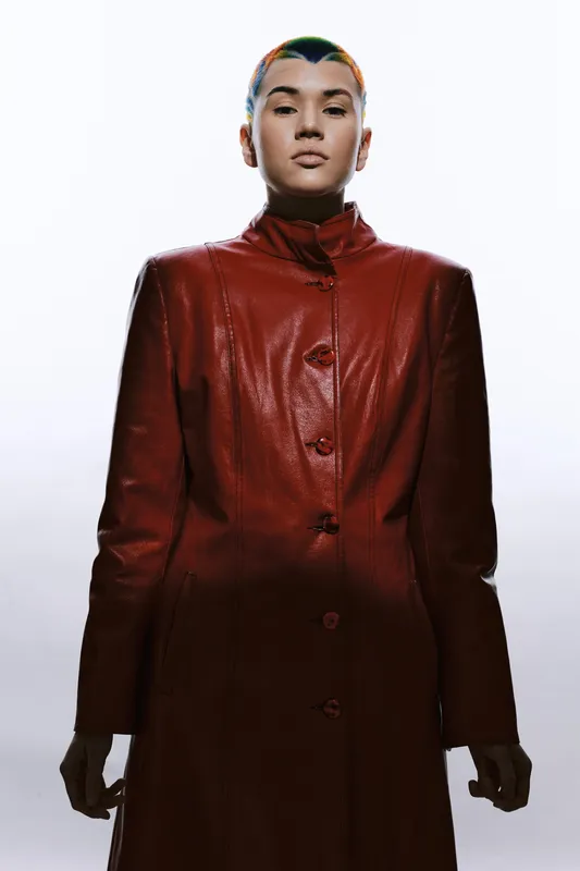 Woman with buzz cut wearing a long red leather coat stands confidently against white background, showcasing modern fashion and strong attitude in studio lighting.