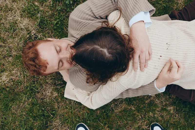 Cheerful couple outdoors, lying on grass, embracing softly, sunlit moment, casual sweaters, intimate connection, natural colors, relaxed mood, warm breeze, park scene