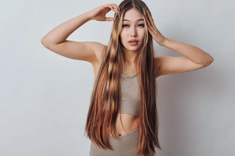 Young woman with long straight hair and casual top posing with hands near head against plain background, fashion and beauty concept with natural makeup.