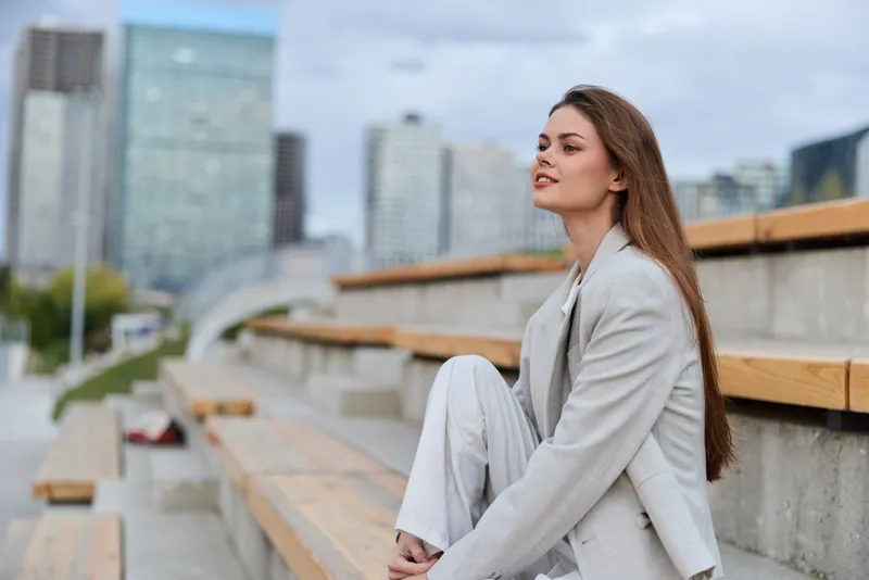 Confident woman in formal attire sitting outdoors against a modern cityscape, showcasing a professional image of business elegance and ambition.