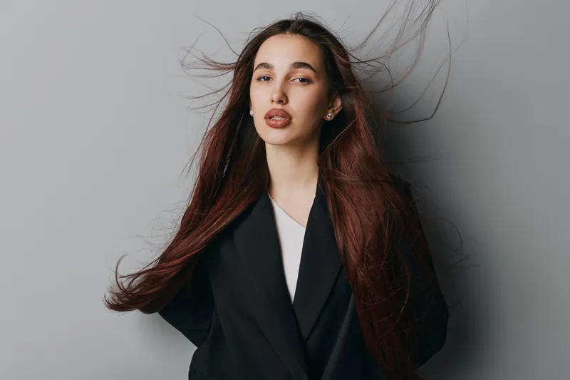Young woman with long flowing hair wearing black blazer stands against gray background with confident expression and natural makeup for fashion portrait concept.