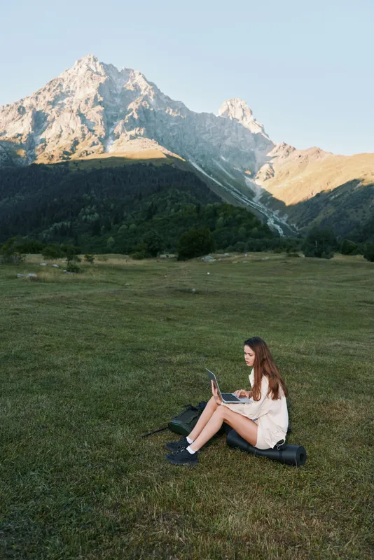 A young woman sits on a grassy meadow at sunset, reading a tablet with a calm, focused expression, surrounded by towering mountains and vast open sky.