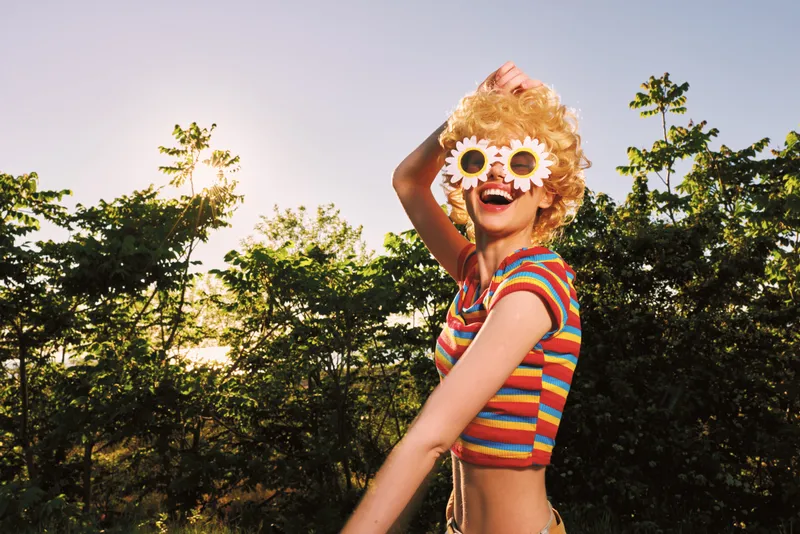 Joyful woman outdoors in retro film colors, wearing a striped top and sunflower sunglasses, lively mood, carefree style, and vibrant sunny atmosphere.