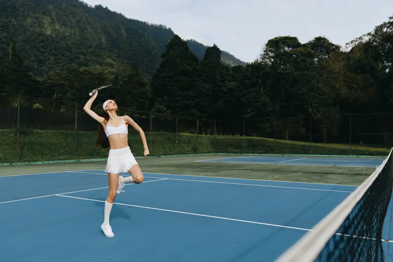 Athletic young woman playing tennis on outdoor court surrounded by lush greenery, showcasing determination and physical activity, wearing a white sports outfit