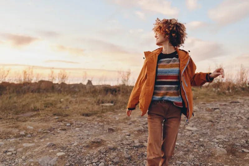 Woman lifestyle outdoors walking on rocky path at sunset, curly hair, brown jacket, striped sweater, joyful relaxed mood, golden light, open sky, rural nature background