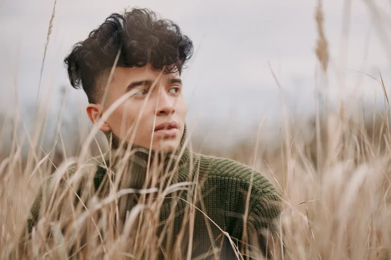 A young man among tall grasses gazes with a quiet, authentic expression that captures a sincere moment and natural atmosphere in soft, warm light.
