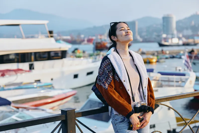 Woman enjoying seaside breeze in a textured coat and scarf by a marina, bright sunlight, distant hills, and boats, creating a stylish travel mood and relaxed lifestyle scene